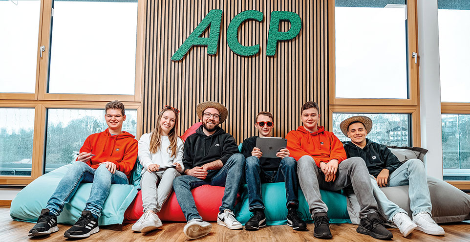 Sechs Personen sitzen auf Sitzsäcken vor einer Fensterfront auf dem Boden. Sie lächeln in die Kamera und tragen teilweise eine Sonnenbrille oder einen Hut. Darüber ist der Schriftzug ACP zu lesen.