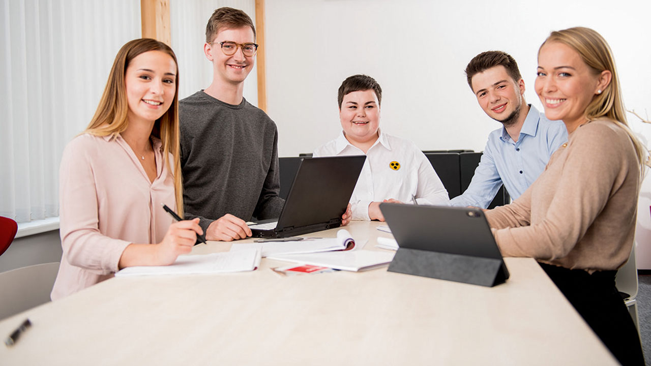 Fünf junge Menschen sitzen an einem Tisch und lächeln in die Kamera