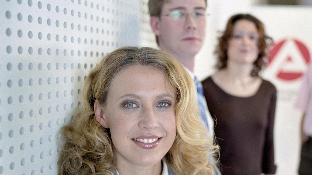 Porträt einer jungen Frau, die freundlich in die Kamera lächelt. Im Hintergrund zwei weitere Personen und das Logo der Agentur für Arbeit.