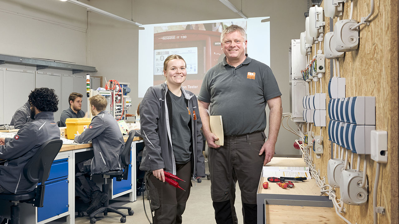 Junge und ältere Person in Arbeitskleidung lächeln in die Kamera, im Hintergrund sitzen weitere Personen