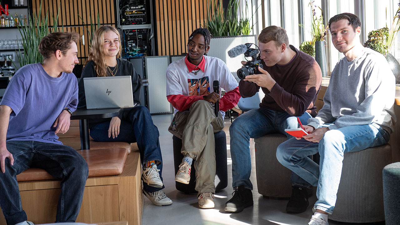 Fünf junge Menschen mit Laptop, Kamera und Mikrofon sitzen in einem Raum
