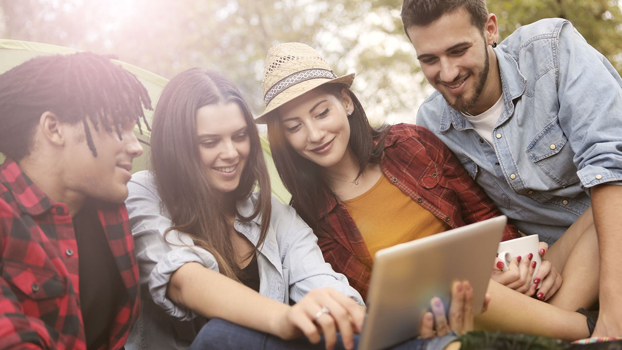 Vier junge Menschen schauen gemeinsam auf ein Tablet, im Hintergrund Bäume und Sonnenstrahlen