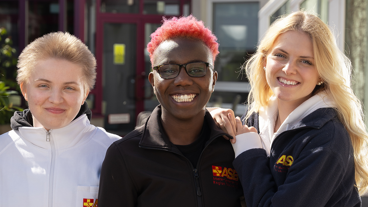 3 junge Mitarbeitende des ASB stehen nebeneinander und lächeln freundlich.