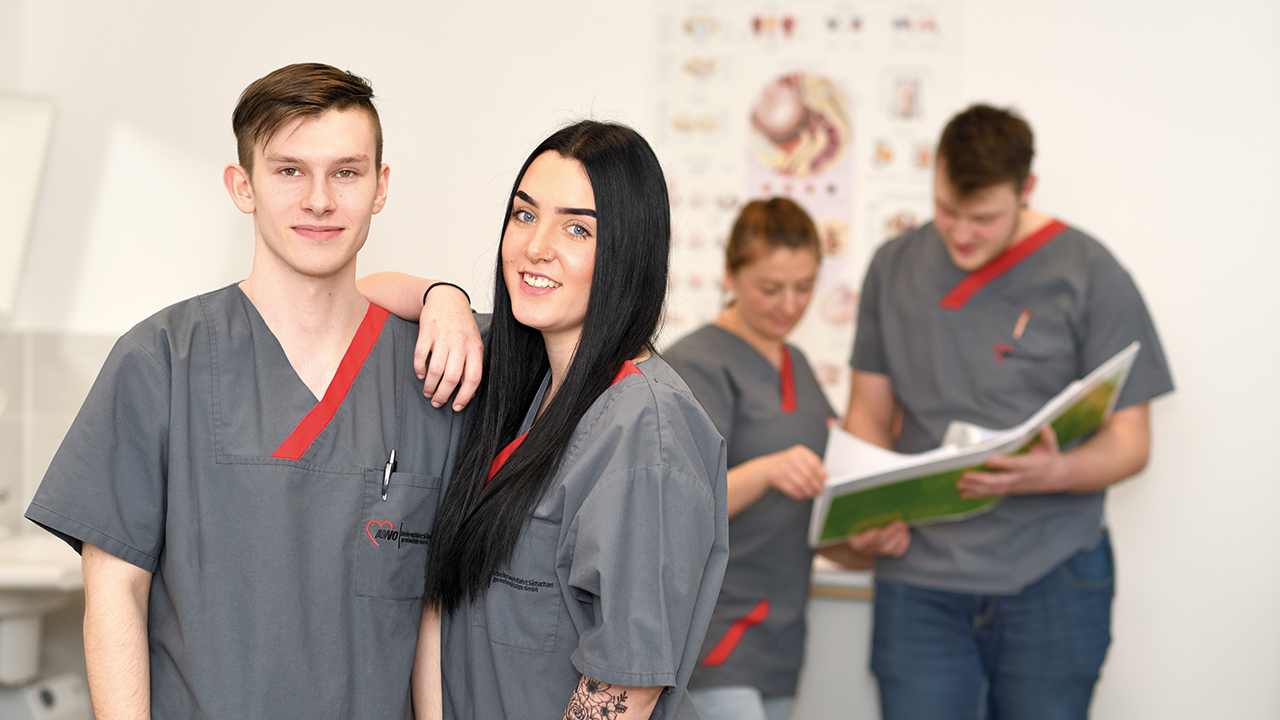Im Vordergrund eine junge Frau und ein junger Mann in Pflegerkleidung, die an einander gelehnt stehen, im Hintergrund eine weitere junge Frau und ein junger Mann, die vor einem anatomischen Poster stehen und in einem Hefter blättern.