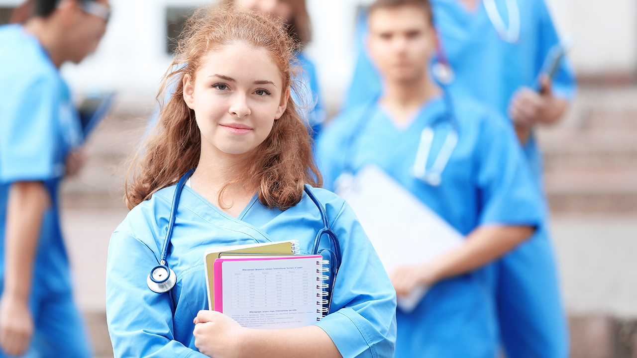 Junge Frau in Arztkleidung hält Unterlagen im Arm und lächelt in die Kamera