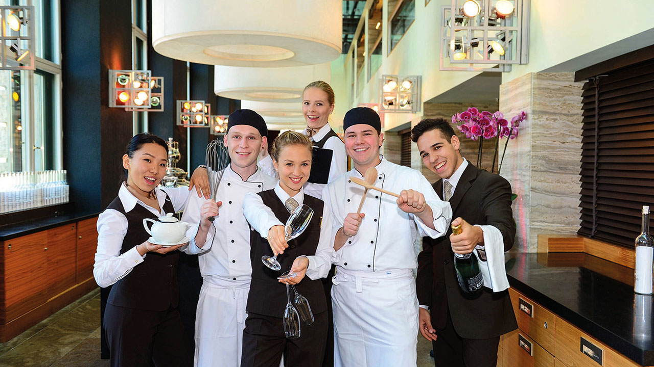 Das Bild zeigt eine Personengruppe die lächelnd in die Kamera blicken. Die Personen tragen verschiedene, in der Gastronomie typische Kleidung.