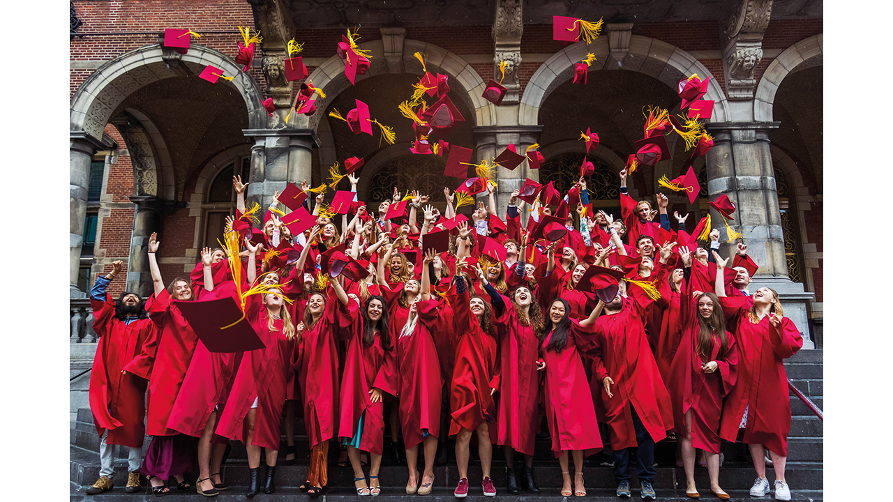 Das Bild zeigt eine Gruppe von Absolventen, die ihren Abschluss feiern. Sie tragen rote Talare und Hüte, und viele werfen ihre Hüte freudig in die Luft. Das Foto fängt einen Moment der Begeisterung und des Erfolgs vor einem historischen Gebäude mit Bögen ein. Die Absolventen lächeln alle und scheinen bester Laune zu sein.