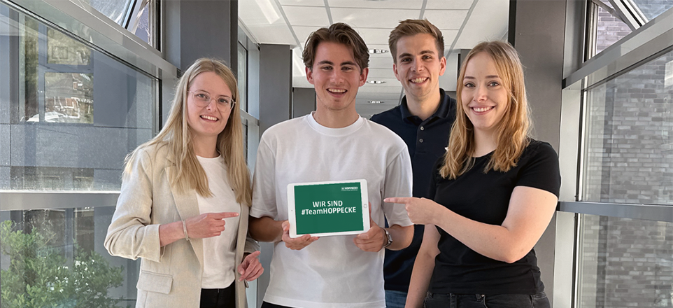 4 junge Menschen in einem Bürogebäude im Halbkörperprofil mit einem Tablet in der Hand auf dem der Spruch Wir sind #TeamHOPPECKE steht