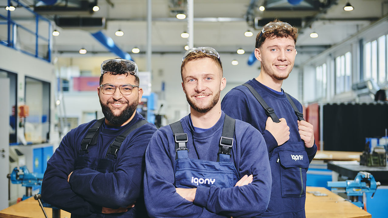 Drei Personen tragen Arbeitskleidung (blauer Pullover und blaue Latzhose) und lächeln in die Kamera. Im Hintergrund ist eine Werkstatt zu sehen.