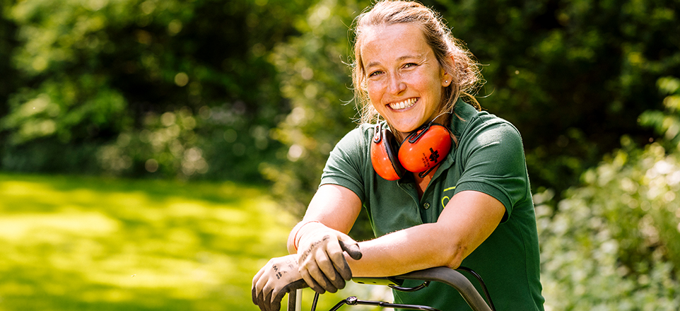 Ein e Person lächelt in die Kamera, sie trägt ein grünes T-Shirt und einen orangenen Gehörschutz. 