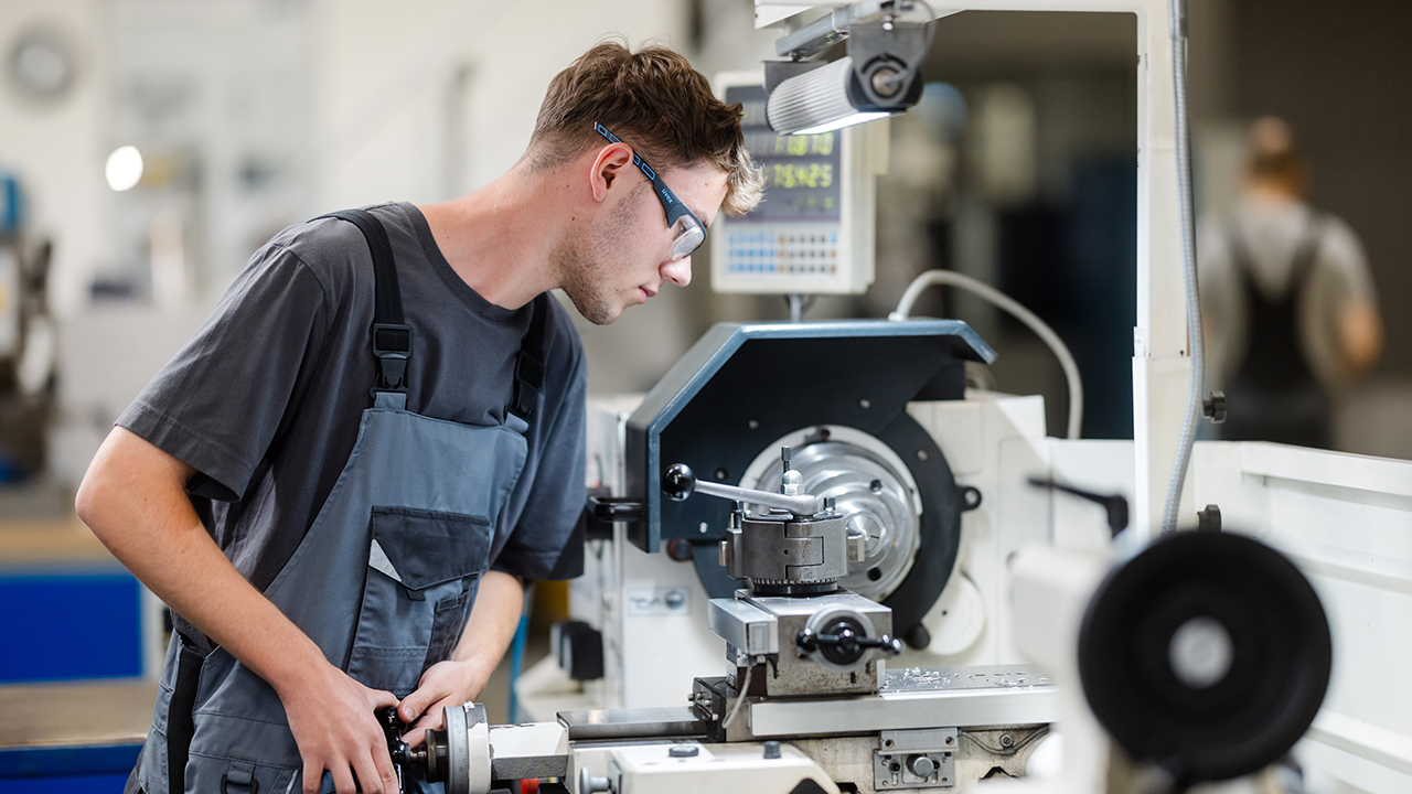 Eine Person arbeitet in einer Werkstatt an einer Maschine, vermutlich einer Dreh- oder Fräsmaschine. Sie trägt Schutzbrille und Arbeitskleidung und bedient konzentriert die Bedienelemente, während sie das Werkstück im Blick behält. Im Hintergrund ist eine industrielle Umgebung mit weiteren Maschinen und unscharfen Personen zu erkennen.