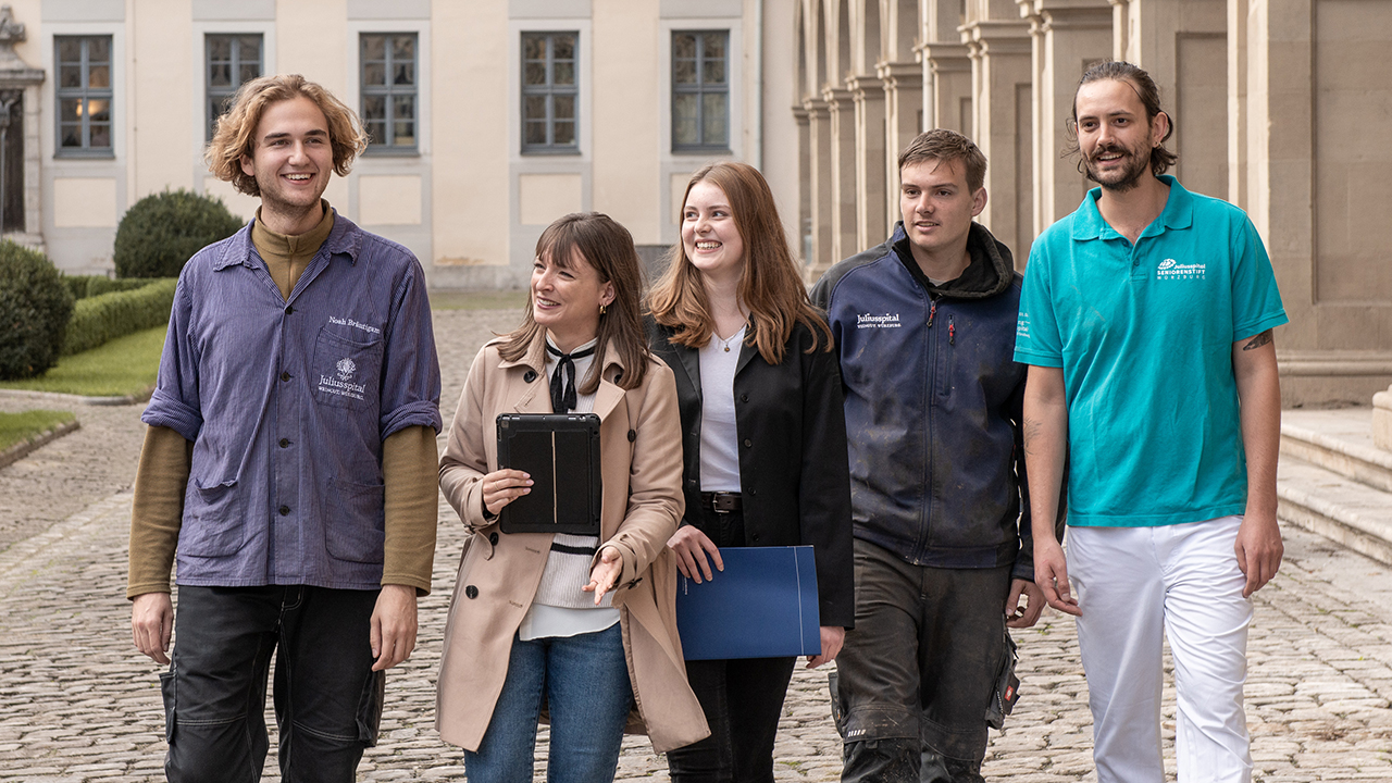 5 junge Menschen laufen nebeneinander im Hof des Juliusspitals. Sie lächeln und wirken gut gelaunt.