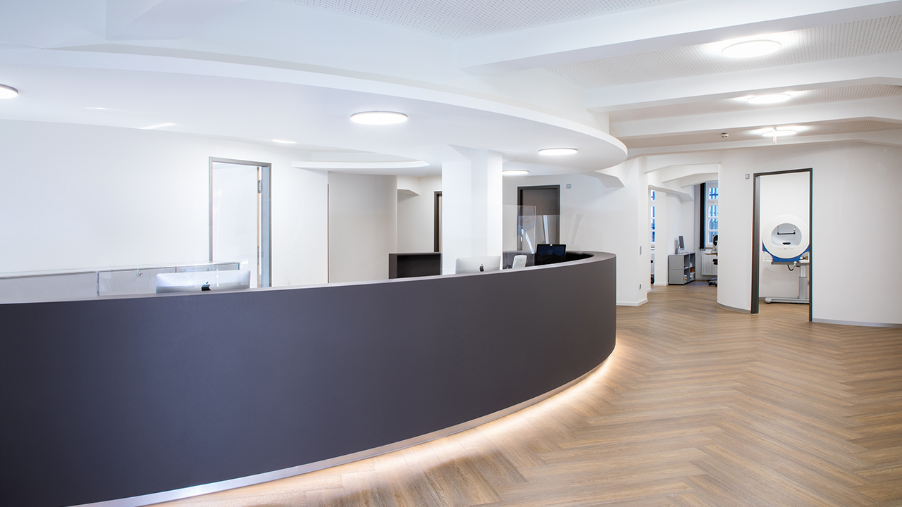 Praxisbild von innen. Hellbeleuchteter Raum mit dunkler Anmeldetheke und Holzboden