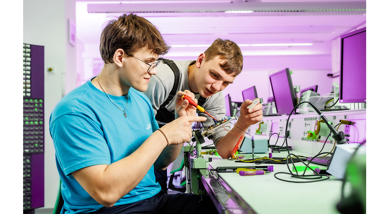 Zwei Menschen sitzen an einem Arbeitsplatz mit Bildschirm und schauen sich ein Kleingerät an