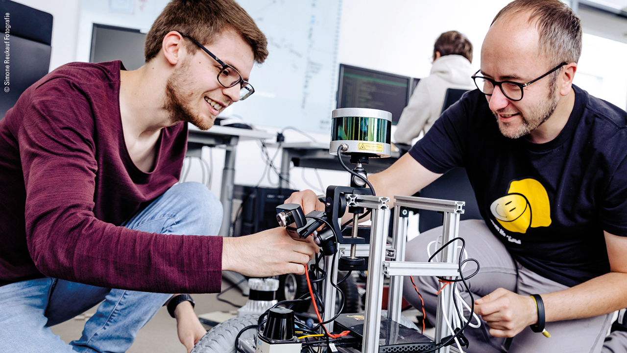 Zwei Personen arbeiten in einem Universitätslabor gemeinsam an einem technischen Gerät. Die Szene zeigt praxisnahes Arbeiten in einem Informatikstudiengang.