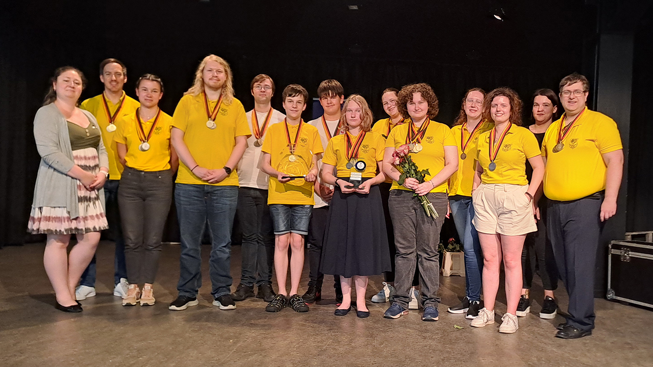 Menschen in gelben Shirts mit Medaillen umhängend. Sie stehen auf der Bühne für ein Gruppenbild.