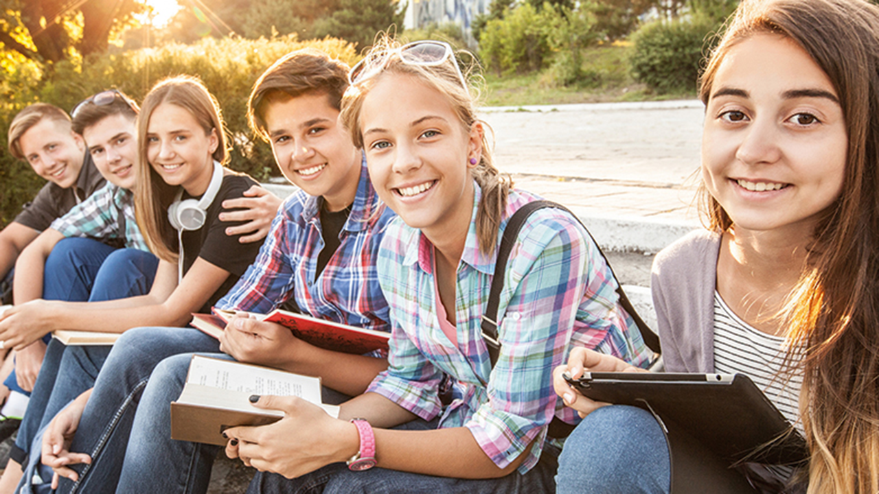 6 junge Menschen sitzen draußen leicht versetzt nebeneinander mit Büchern und Tablet in der Hand. 