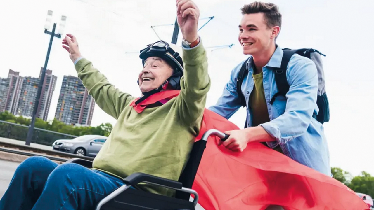 Älterer fröhlicher Herr im Rollstuhl sitzend mit rotem Umhang wird von jungem Mann durch eine Stadt geschoben.