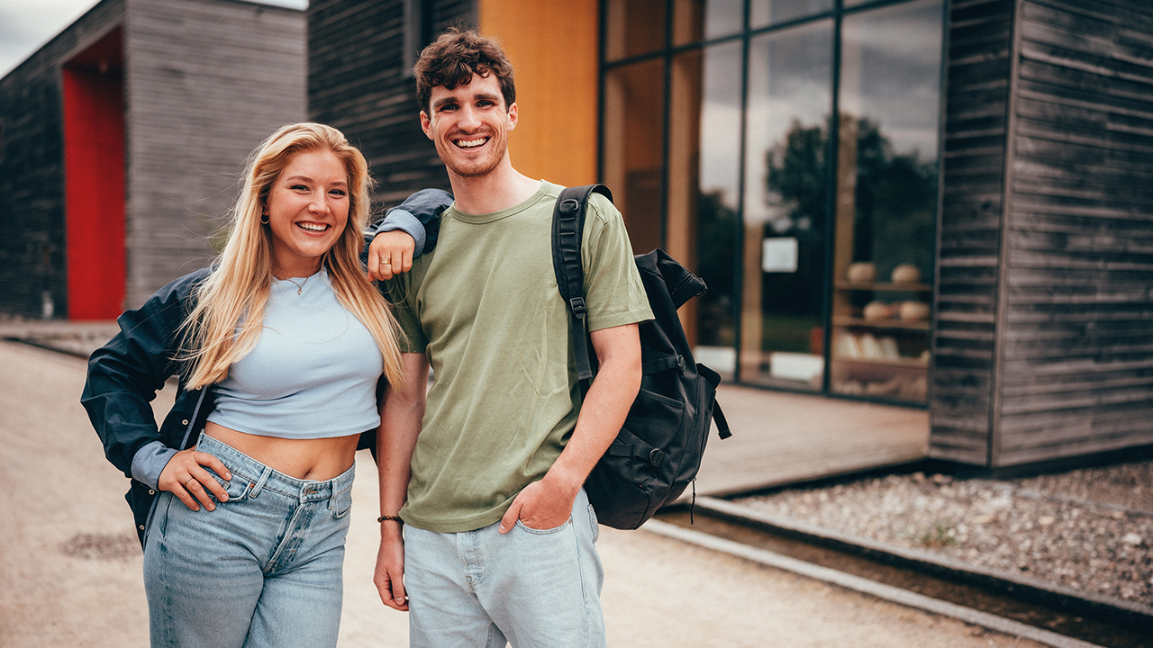 Zwei Studierende lächeln in die Kamera und stehen vor dem Campus