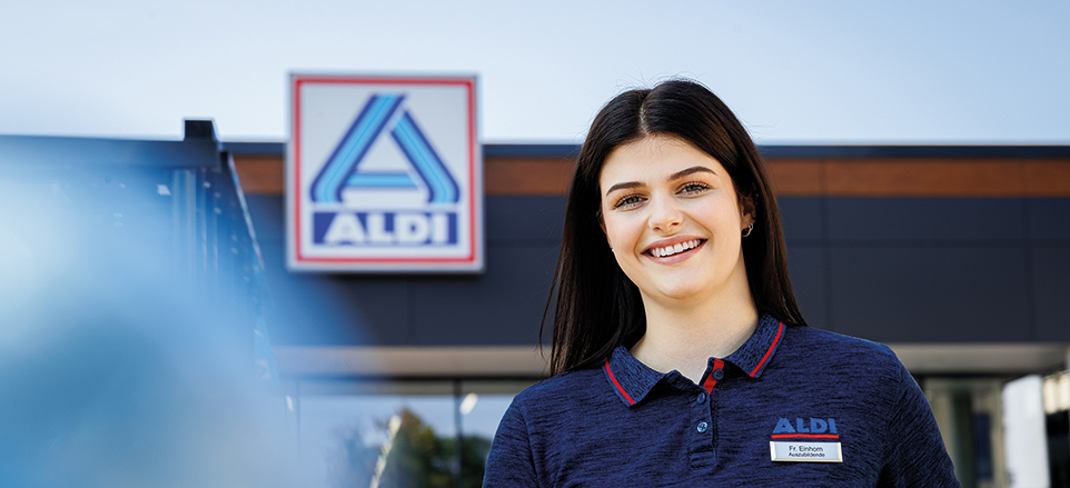 Junge Person im Poloshirt mit Aldi-Logo steht vor einer Aldi-Filiale