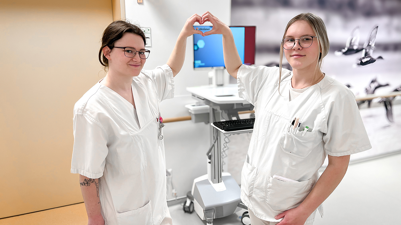 Das Bild zeigt zwei junge Frauen in einem Krankenhaus. Beide tragen helle Arbeitskleidung und stehen nebeneinander. Sie lächeln leicht und formen mit ihren erhobenen Armen gemeinsam ein Herzsymbol über ihren Köpfen.

Im Hintergrund ist medizinische Ausstattung zu sehen, darunter ein Gerät mit Bildschirm auf einem rollbaren Wagen. Die Umgebung wirkt sauber und modern. Insgesamt vermittelt das Bild einen freundlichen, kollegialen Eindruck und könnte Teamarbeit oder Zusammenhalt im Gesundheitswesen symbolisieren.