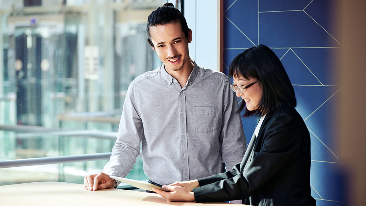 Eine junge lächelnde Person zeigt einer anderen jungen lächelnden Person etwas auf dem Tablet