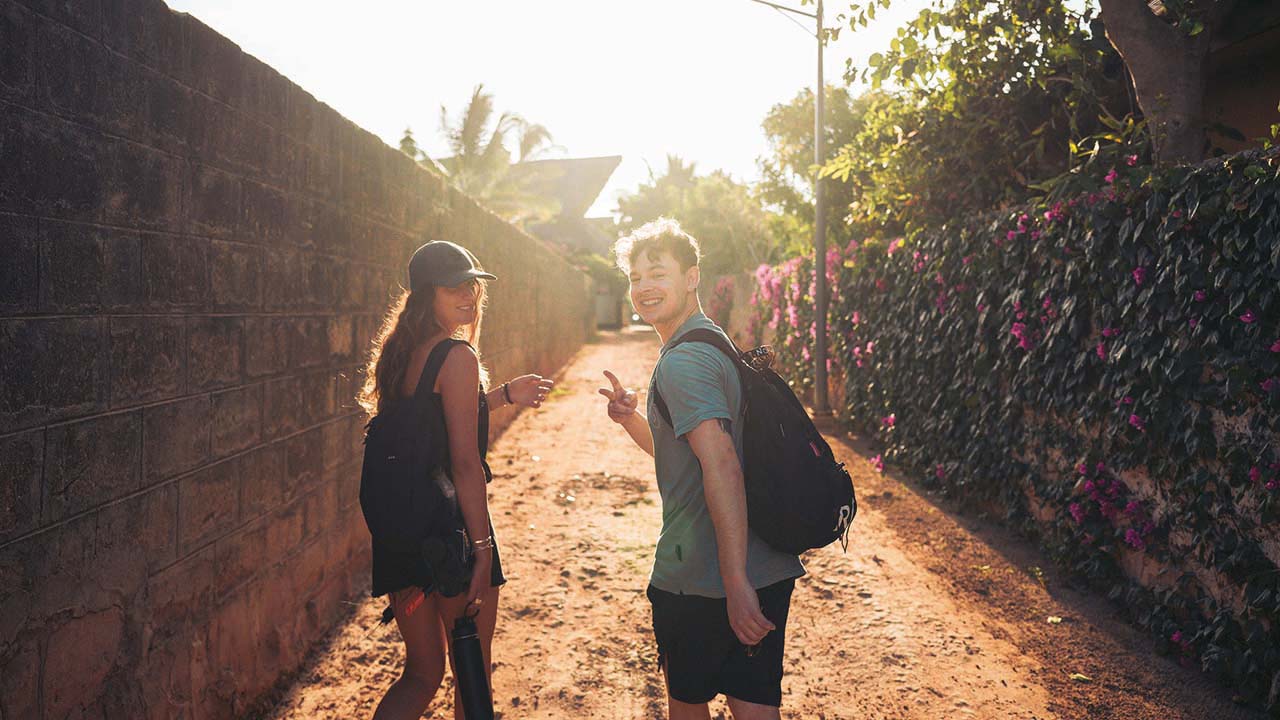 Zwei junge Menschen laufen Sandstraße entlang und schauen rückswärts in die Kamera