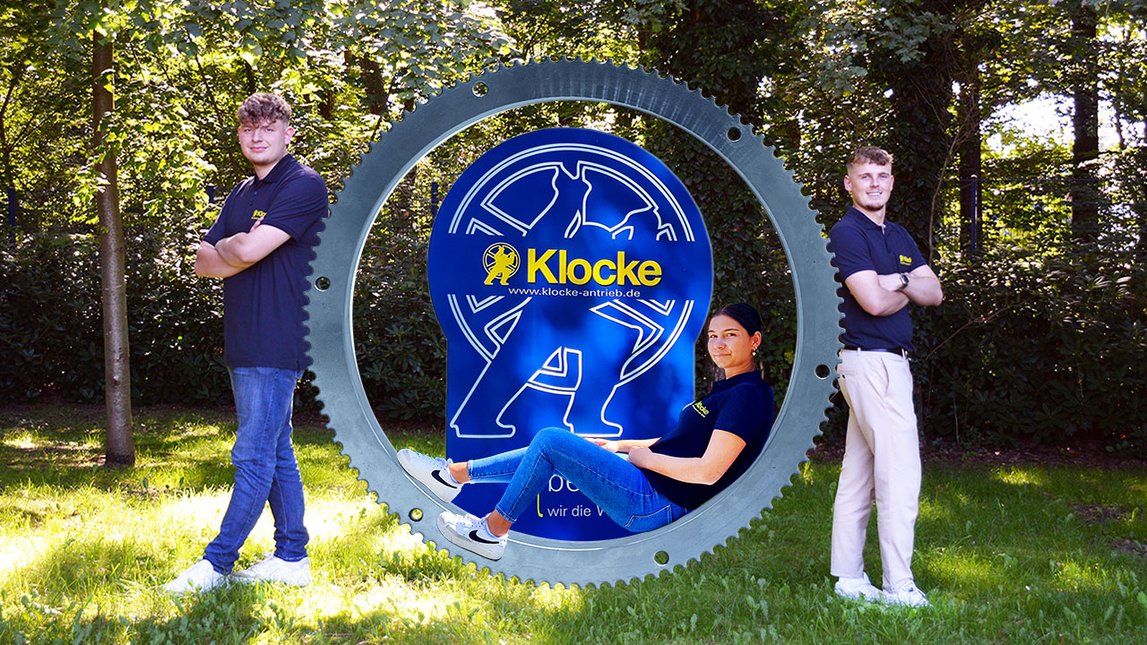 Auf dem Bild sind drei Personen zu sehen, zwei stehen mit dem Rücken an ein großes Zahnrad gelehnt, die dritte sitzt in dem Zahnrad. Im Hintergrund ist das Logo der Firma zu sehen.