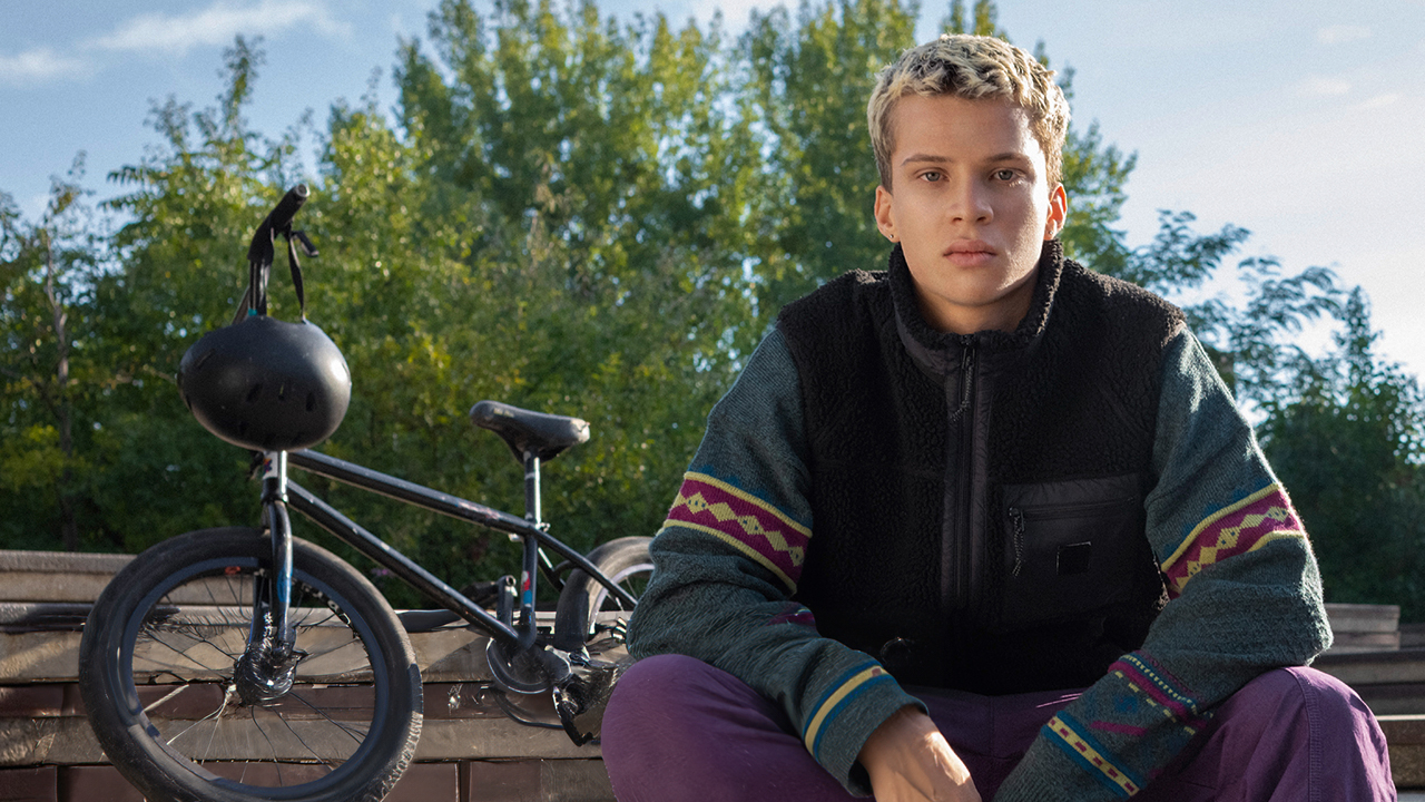 Ein junger Mann sitzt auf einer Treppe. Im Hintergrund liegt ein Rad un einige Bäume sind zu sehen.