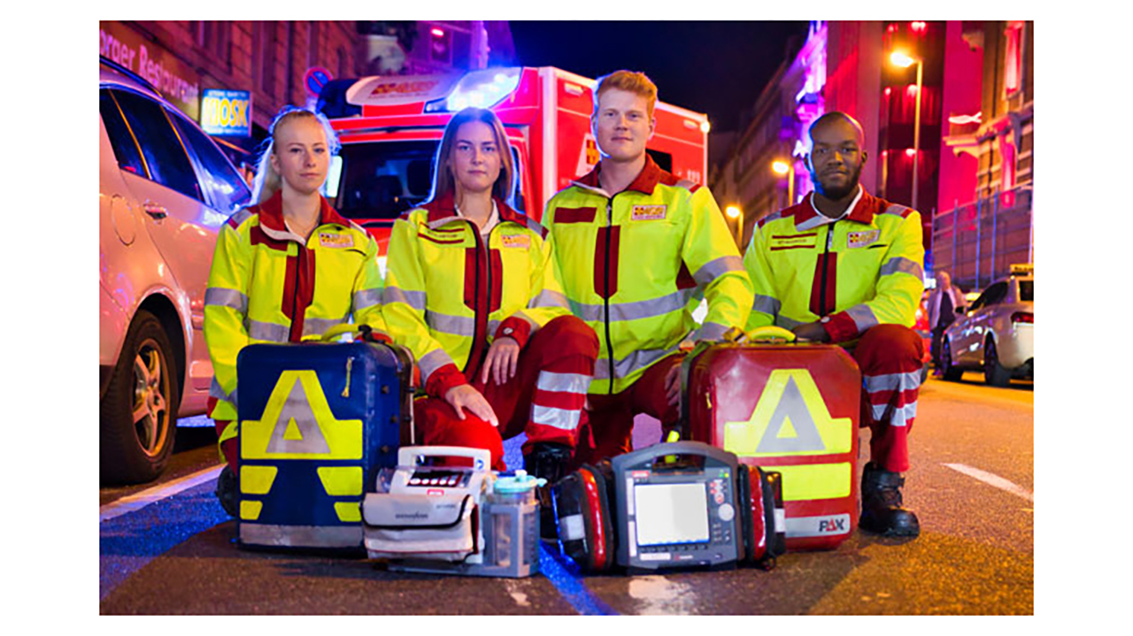 Gruppenbild von Sanitätern mit Ausrüstung vor einem Einsatzauto auf der Straße. Es ist dunkel. Die Lichter spiegeln sich.