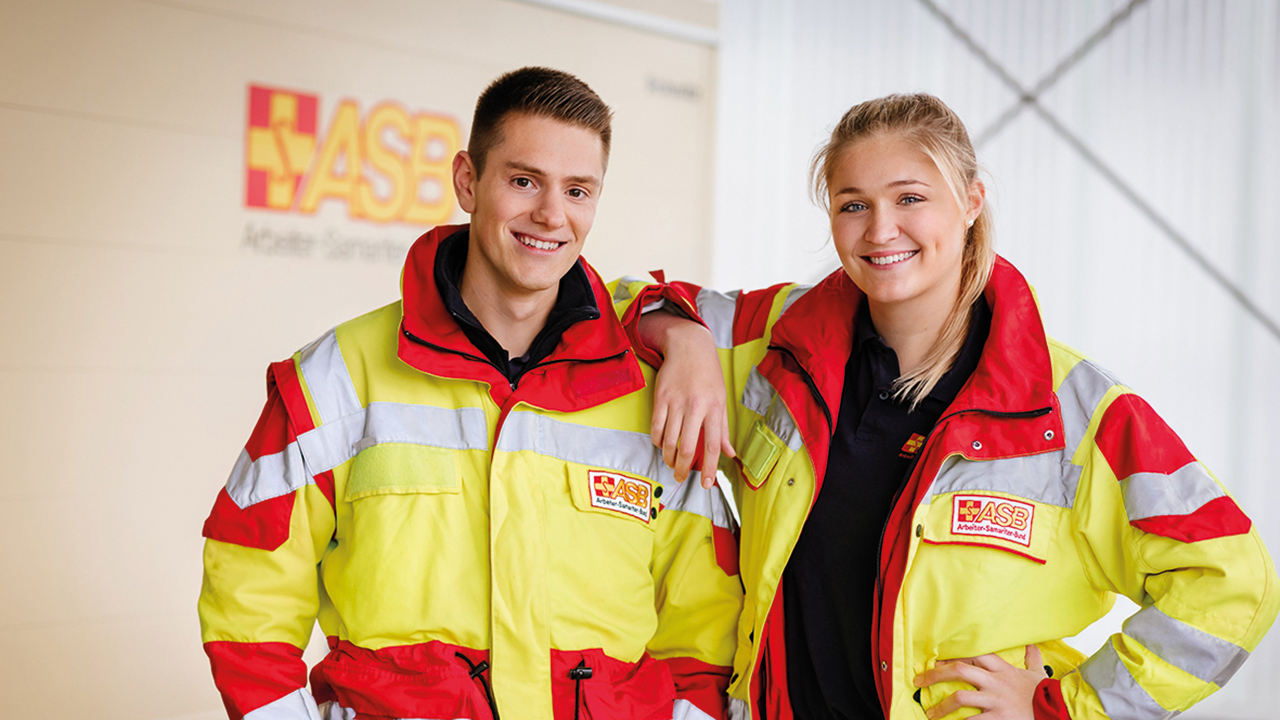 Ein junger Mann und eine junge Frau stehen nebeneinander in der gelb roten Arbeitskleidung des ASB. Im Hintergrund sieht man das ASB Logo.