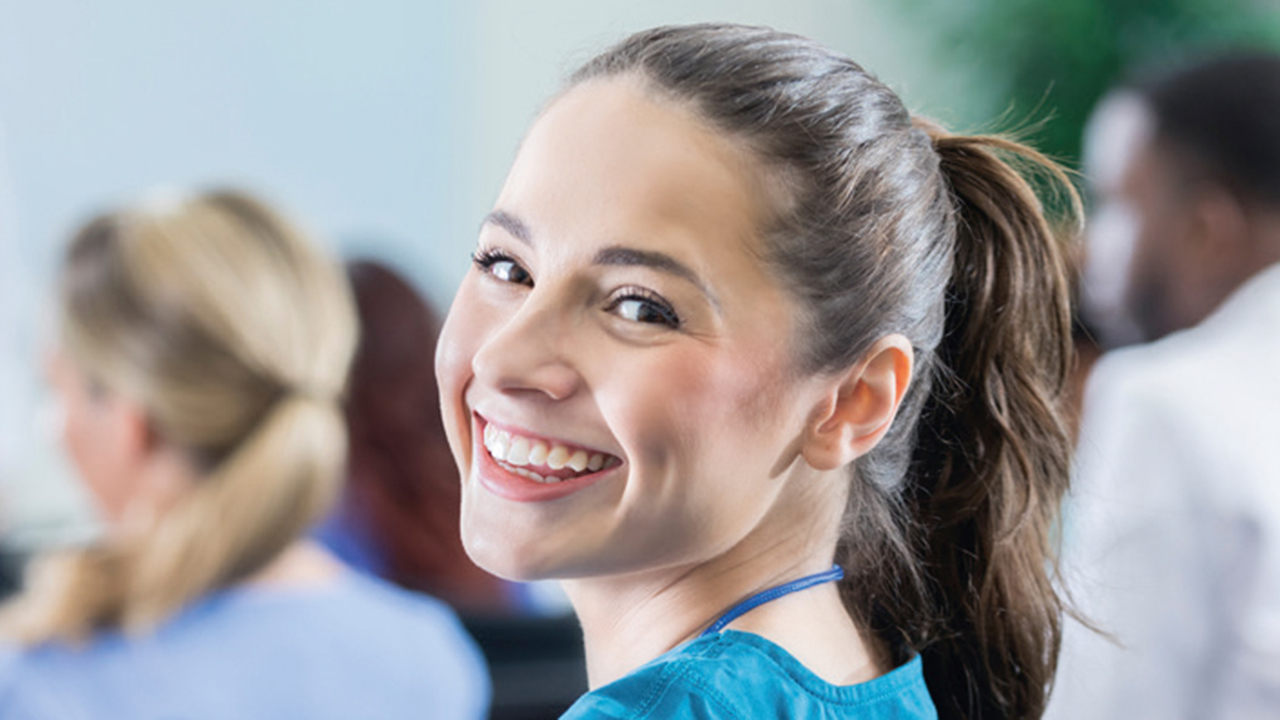 Junge Person im Unterrichtsraum dreht sich lächelnd zur Kamera um, im Hintergrund sitzen weitere Personen