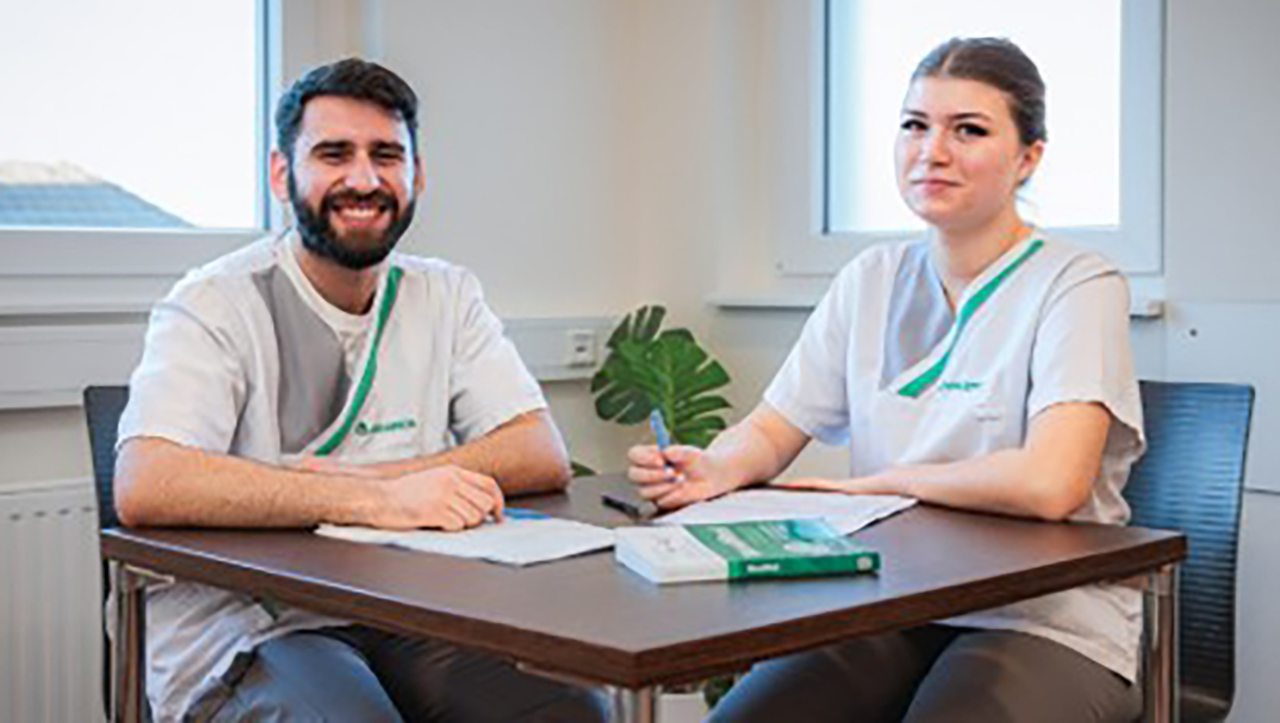 Zwei Menschen sitzen an einem Tisch und schauen in die Kamera