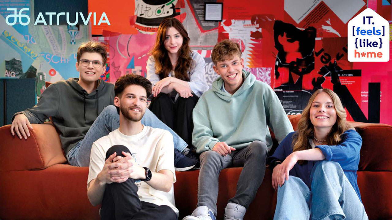 Fünf Personen sitzen auf einer Couch und lächeln in die Kamera. Rechtsseitig der Spruch "IT. [feels] (like} home"