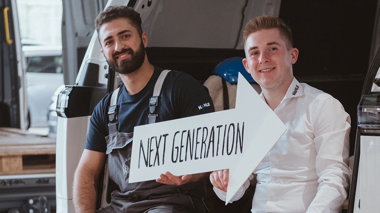 Zwei junge Männer sitzen im geöffneten Kofferraum eines PKWs in Arbeitskleidung und halten ein pfeilförmiges Schild mit der Aufschrift: Next Generation in der Hand. Beide lächeln in die Kamera.