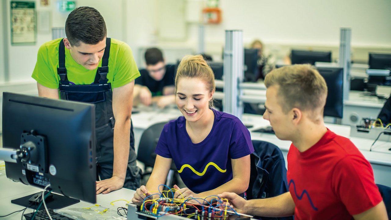 Drei junge Menschen sitzen in einem Computerraum und schauen sich gemeinsam am Schreibtisch etwas an