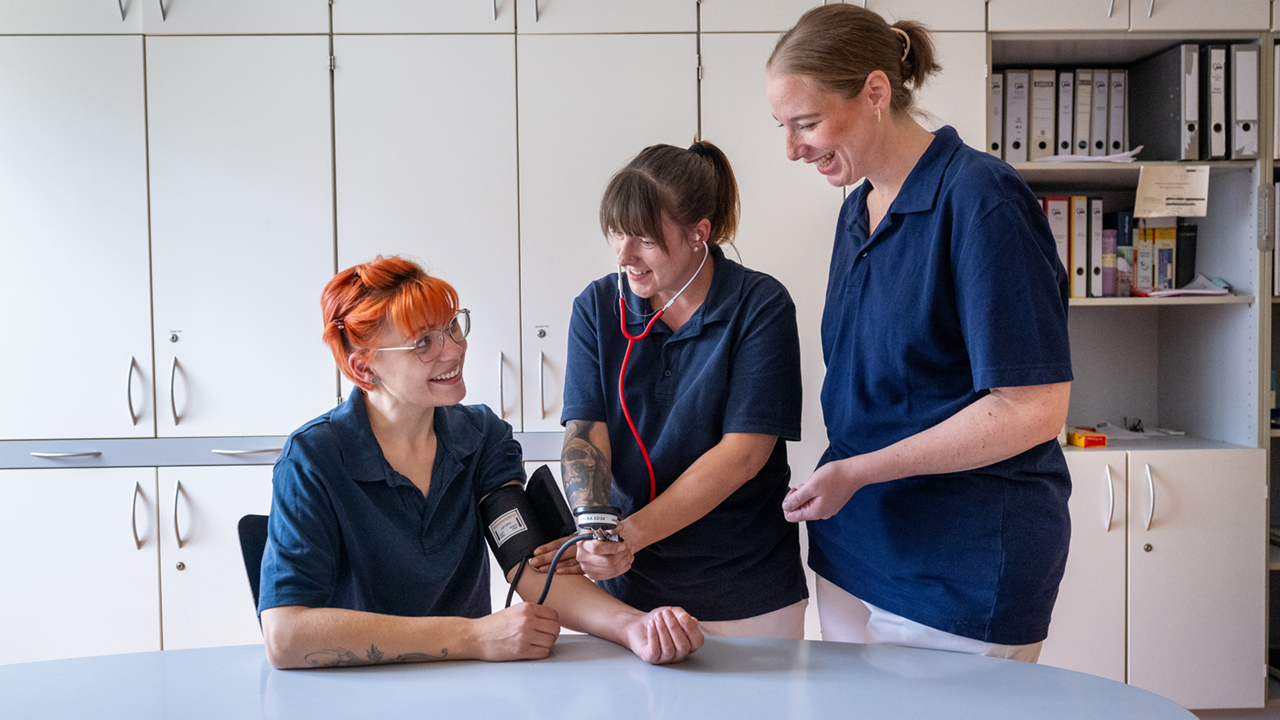 Drei junge lächelnde Personen in blauen Poloshirts, eine Person misst einer sitzenden Person den Blutdruck