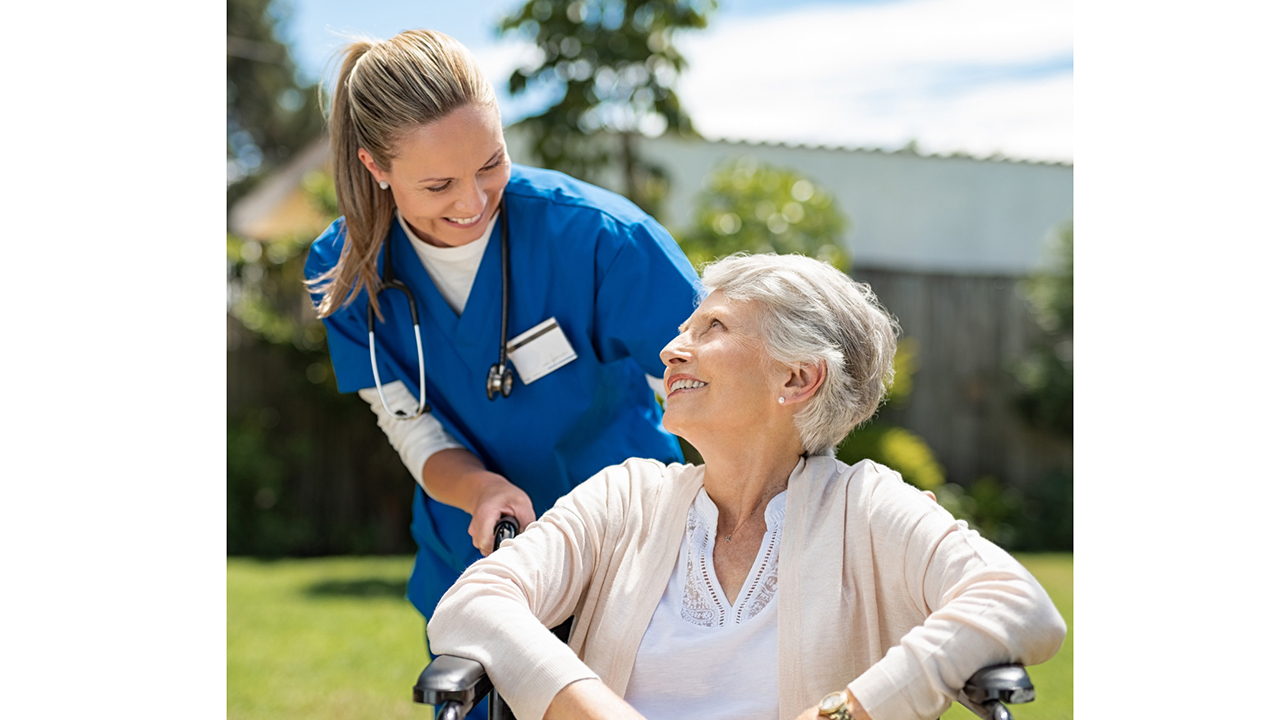 Junge Frau schiebt betagte Dame im Rollstuhl an der frischen Luft. Sie lächeln sich an.