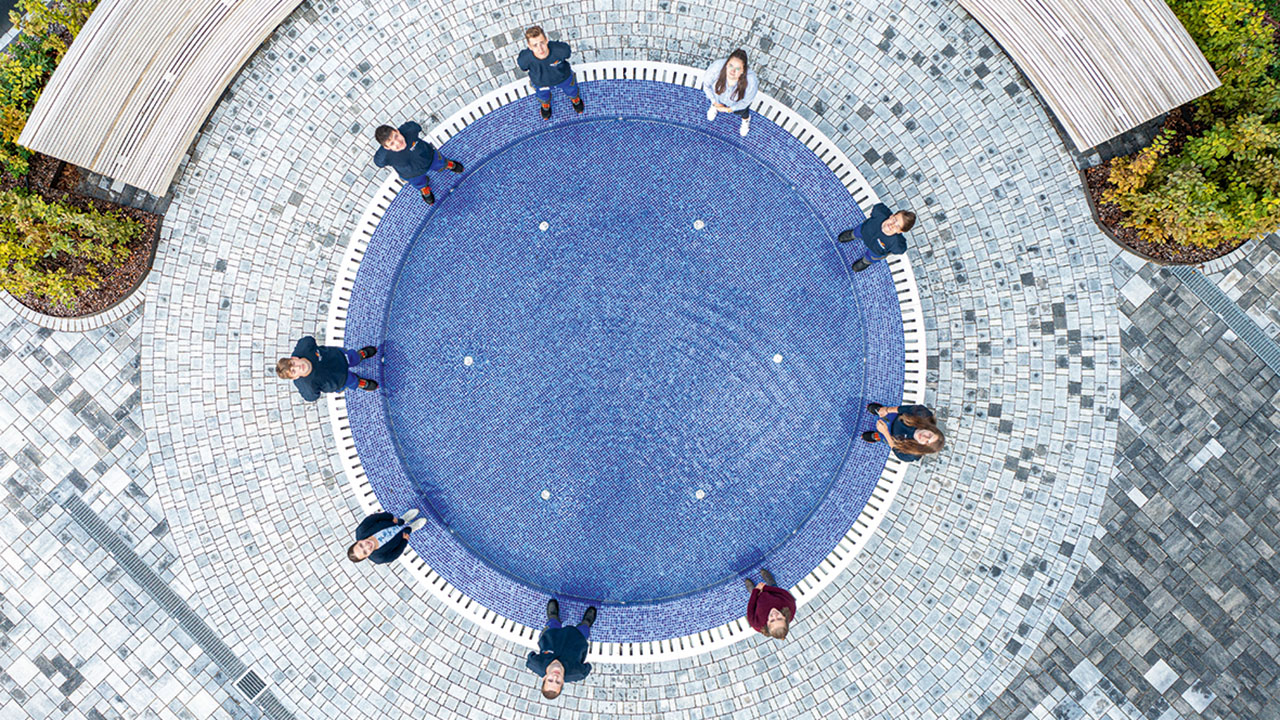 Aufnahme eines runden gefliesten Wasserbeckens von oben, um das neun junge Personen herum stehen 