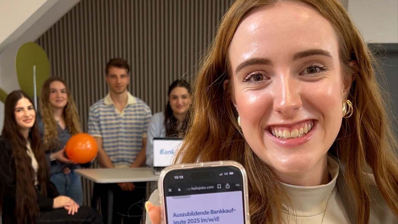 Junge Dame mit langen braunen Haaren streckt lächelnd ihr Handy in die Kamera