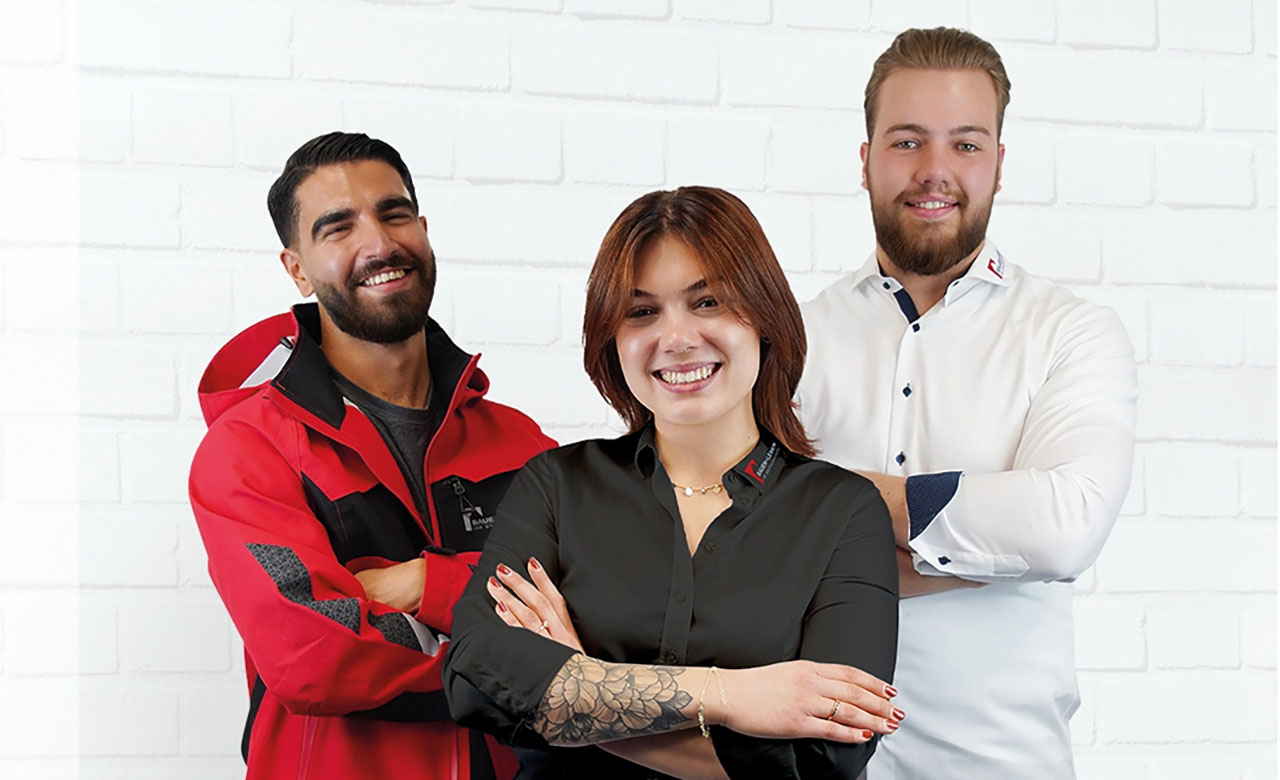 Drei Personen stehen lächelnd vor einer weißen Ziegelwand – eine in roter Jacke, eine in dunklem Hemd mit sichtbarem Tattoo, eine in weißem Hemd.