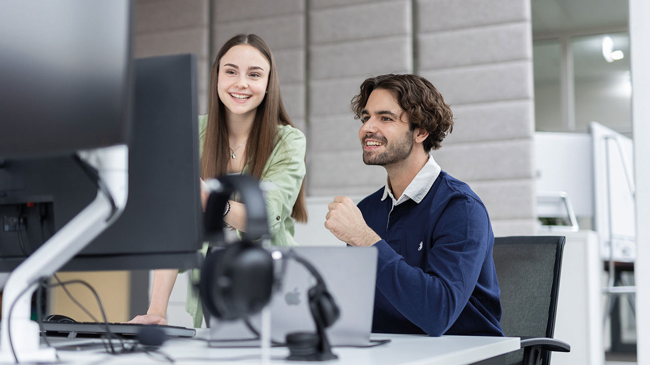 Auf dem Bild sind zwei Personen zu sehen, die auf einen Bildschirm sehen. Im Hintergrund sieht man ein Büro.