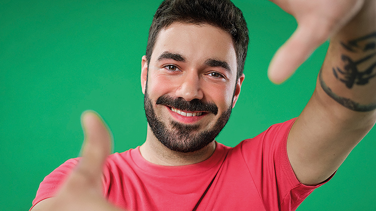 Zu sehen ist eine Person in einem roten T-Shirt vor einem grünen Hintergrund, die ihre Hände vor dem Gesicht zu einem Rahmen hält.