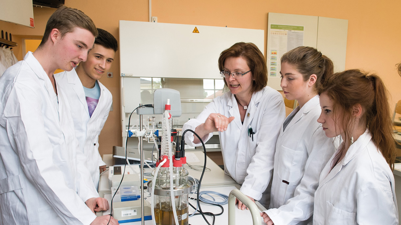 Schülergruppe in einem Chemiekabinett in Schutzkleidung bekommt gerade von einer Lehrerin etwas erklärt.