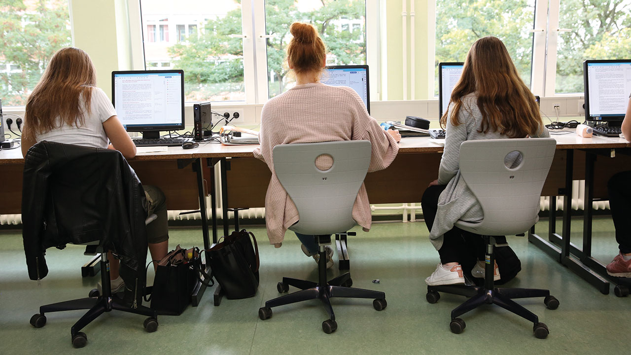 3 Menschen sitzen mit dem Rücken zur Kamera auf Stühlen an einem Computer