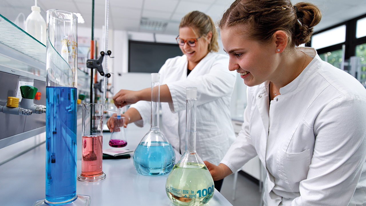 Zwei junge Frauen arbeiten in einem modernen Labor an chemischen Experimenten. Beide tragen weiße Laborkittel. Im Vordergrund steht eine Frau an einem Labortisch und betrachtet konzentriert mehrere Glasgefäße mit farbigen Flüssigkeiten – darunter Blau- und Grüntöne. Im Hintergrund mischt eine weitere Frau eine pinkfarbene Flüssigkeit in einem Kolben. Auf dem Tisch stehen verschiedene Messzylinder, Kolben und Laborgeräte. Der Raum ist hell, sauber und wirkt professionell ausgestattet.