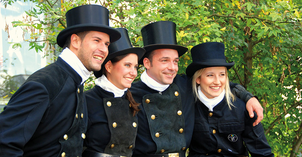 Vier Personen in traditioneller schwarzer Schornsteinfeger-Uniform und Zylinder sitzen lachend nebeneinander im Grünen.