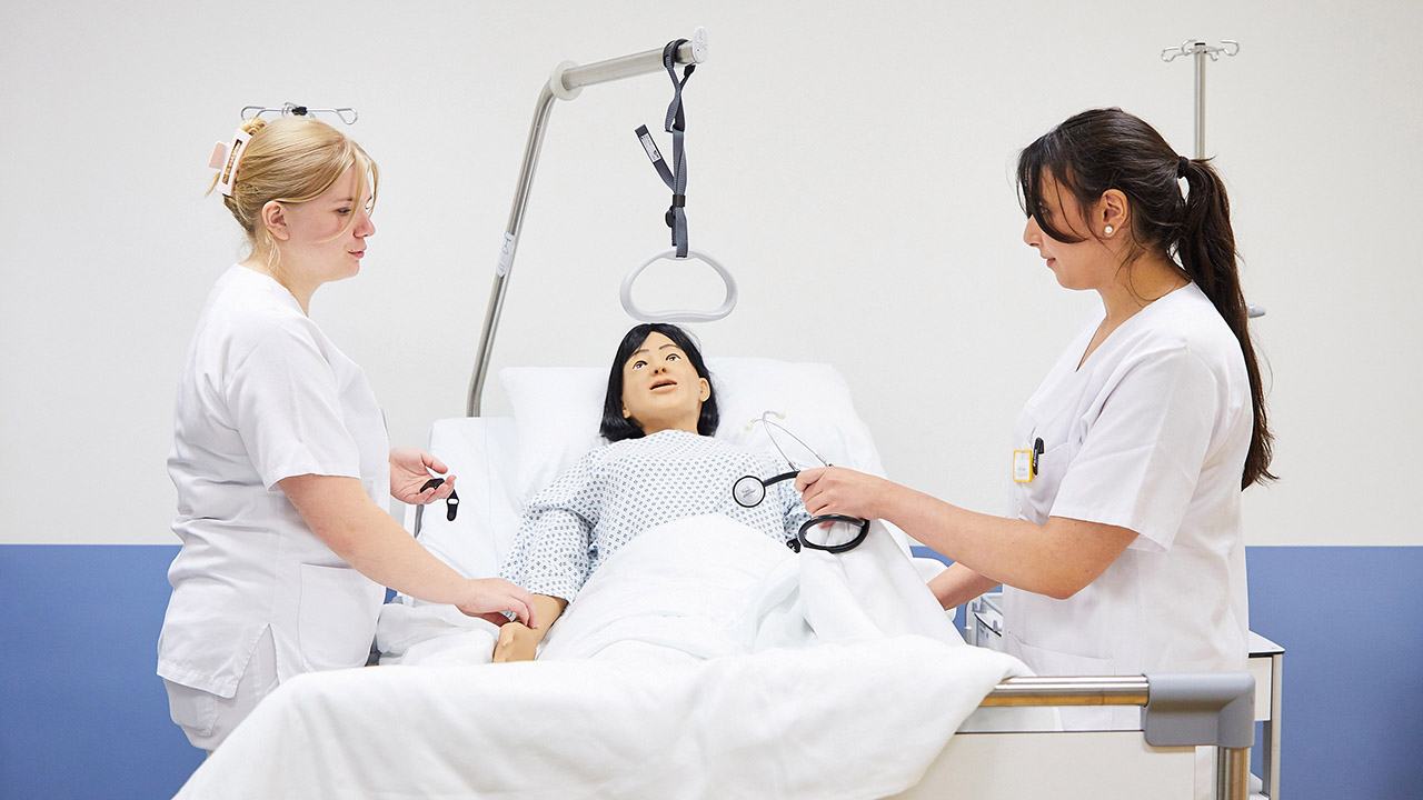 Zwei Mitarbeitende stehen an einem Krankenbett mit Simulationspuppe