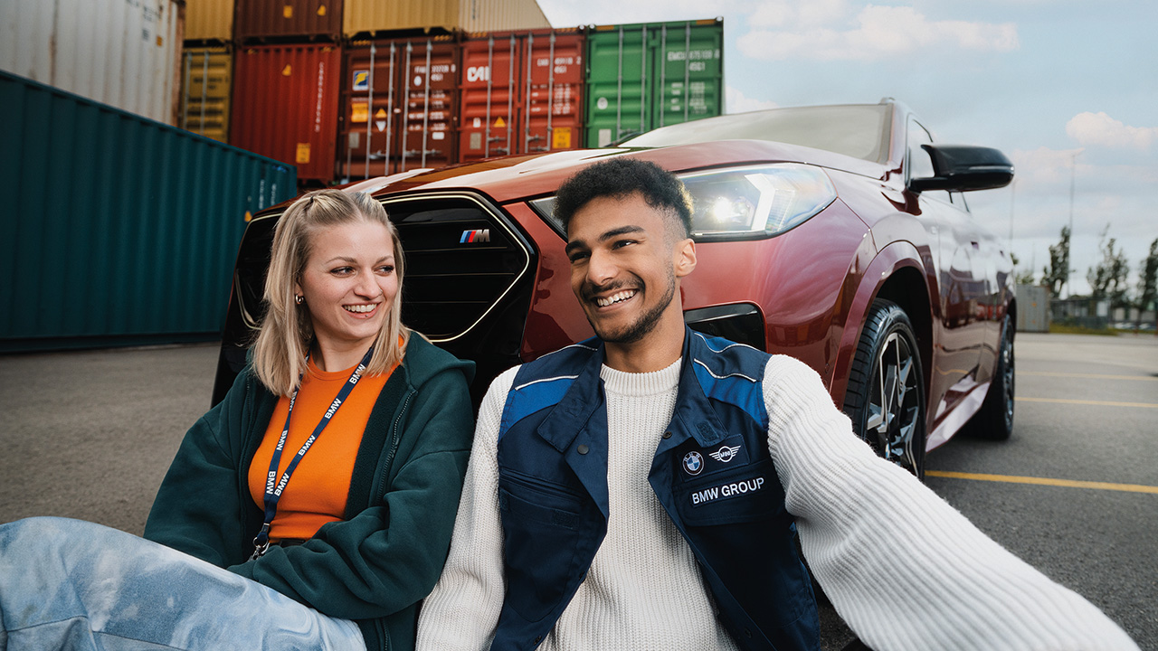 Zwei Menschen sitzen vor einem dunkelroten Auto