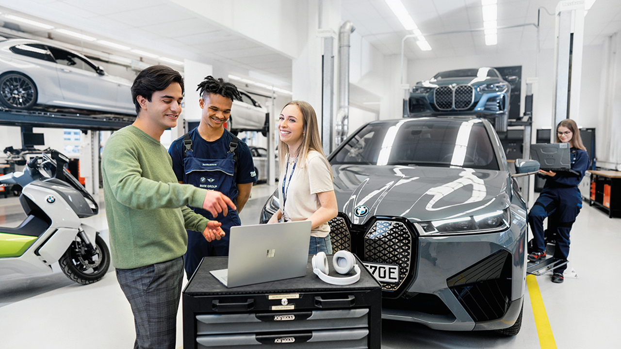 Szene in einer modernen BMW Werkstatt. Man sieht verschiedene Fahrzeuge von BMW, im Vordergrund drei junge Menschen, die an einem Laptop eine Besprechung haben, im Hintergrund eine junge Mitarbeiterin bei der Fahrzeuganalyse mit einem Rechner.